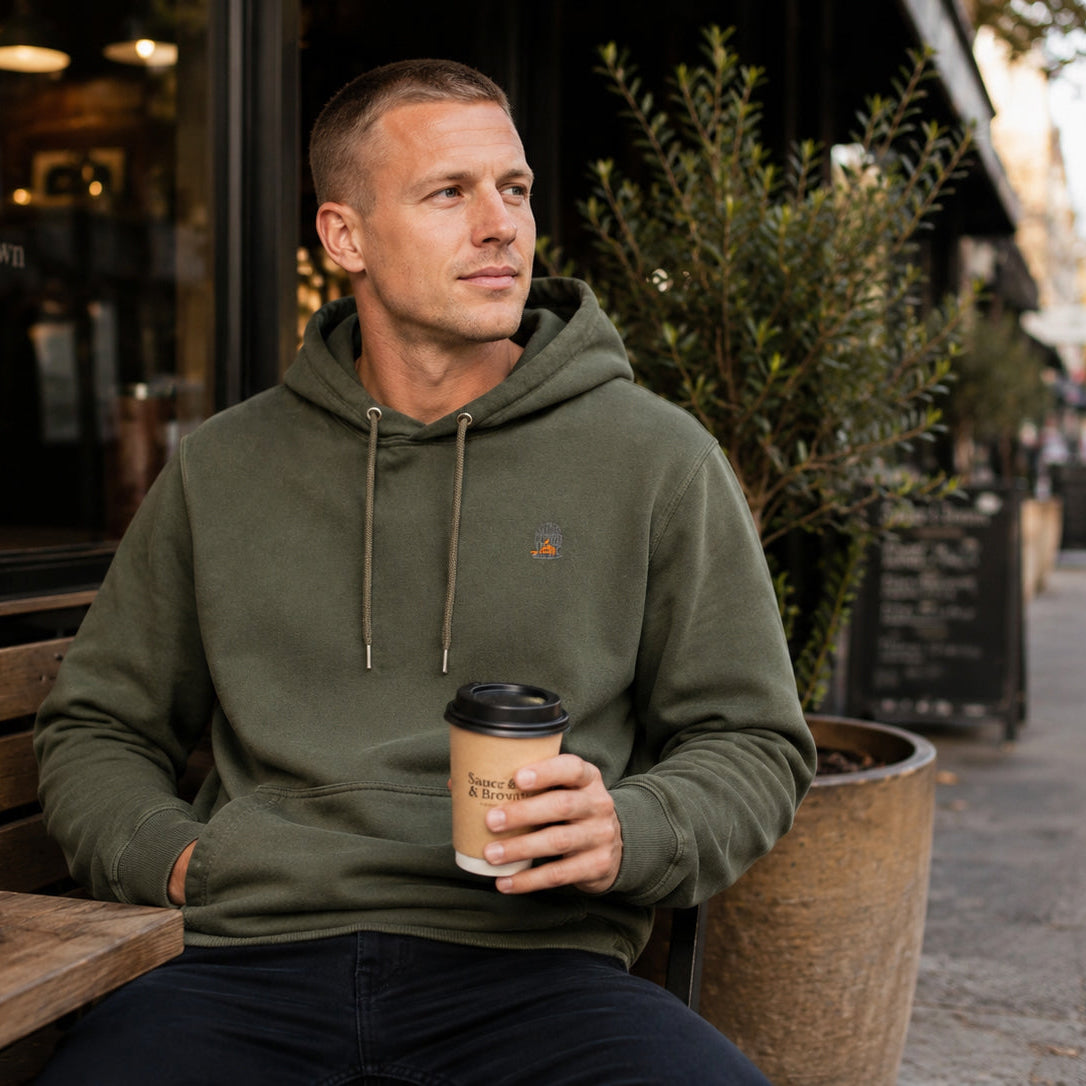 Man wearing the khaki green Tarn hoodie by Sauce & Brown sitting outside a café with a takeaway coffee.