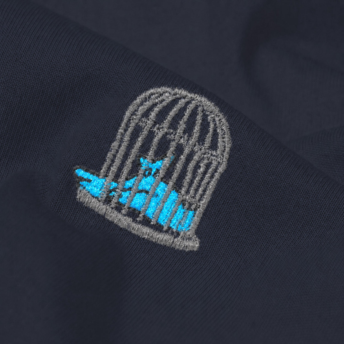 Close-up of the embroidered birdcage logo on the Sauce & Brown Atlantic navy T-Shirt showing the premium cotton fabric.