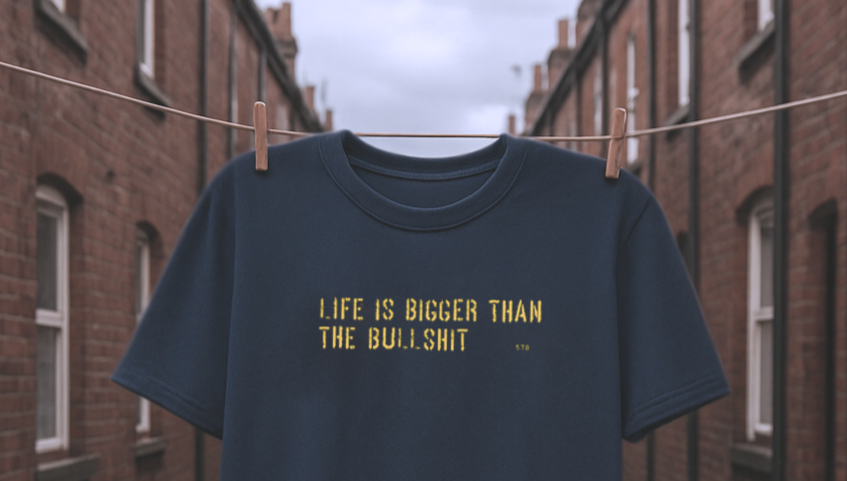 Navy blue t-shirt with yellow text 'LIFE IS BIGGER THAN THE BULLSHIT' on a clothesline against a brick building background.