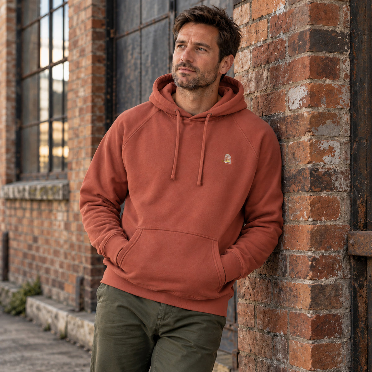 Man wearing the Sauce & Brown Kiln Hoody in Vintage Washed Terracotta, leaning against a weathered brick wall. Relaxed fit with embroidered birdcage logo.