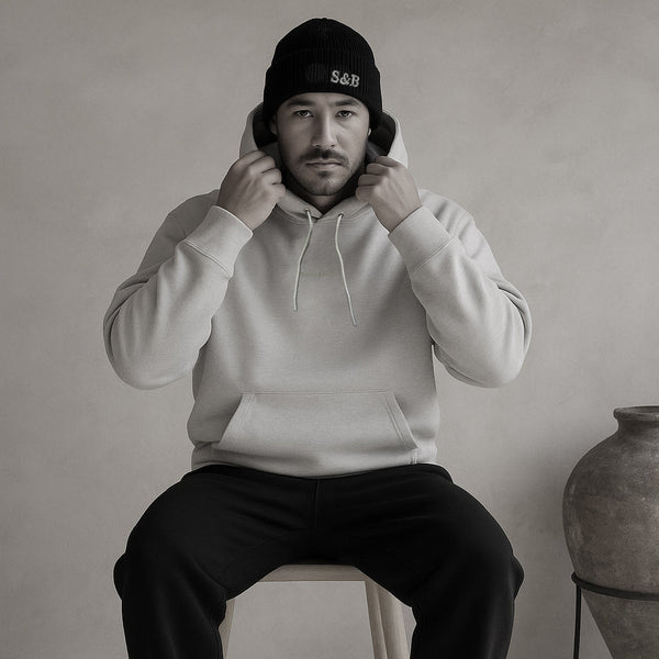 Man wearing a cream hoodie and black beanie sitting on a stool against a plain background