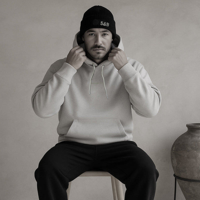 Man wearing a cream hoodie and black beanie sitting on a stool against a plain background