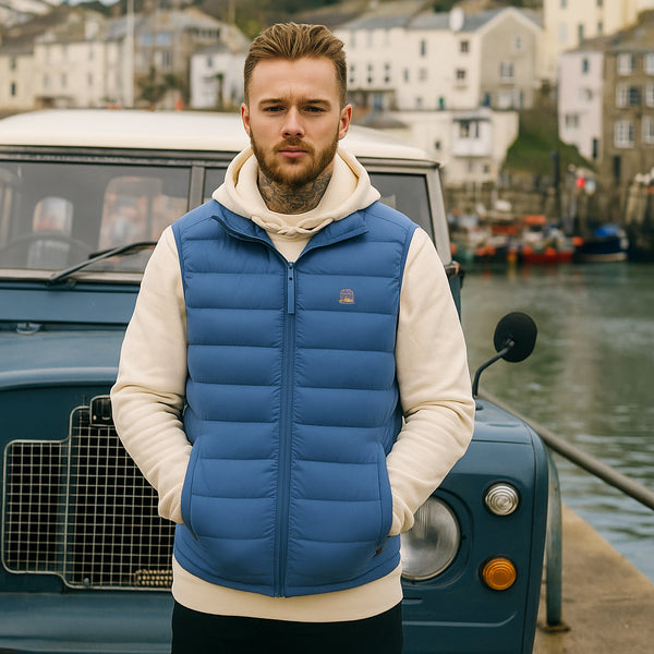 Man wearing a blue puffer vest standing in front of a vintage car by a waterfront.