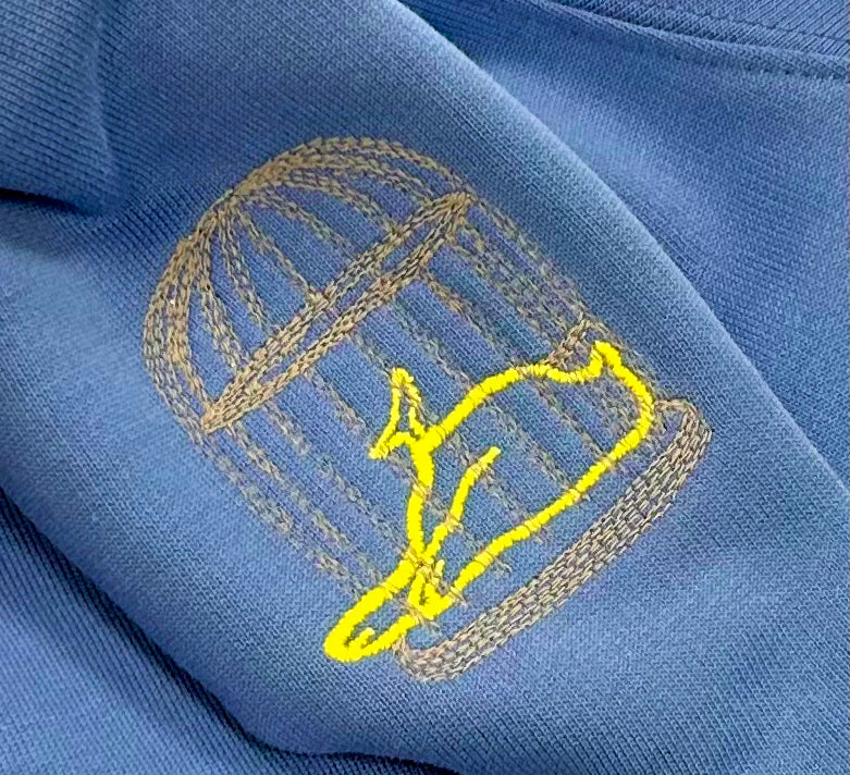 Close-up of yellow bluebird logo embroidery on Sauce & Brown blue crewneck sweatshirt